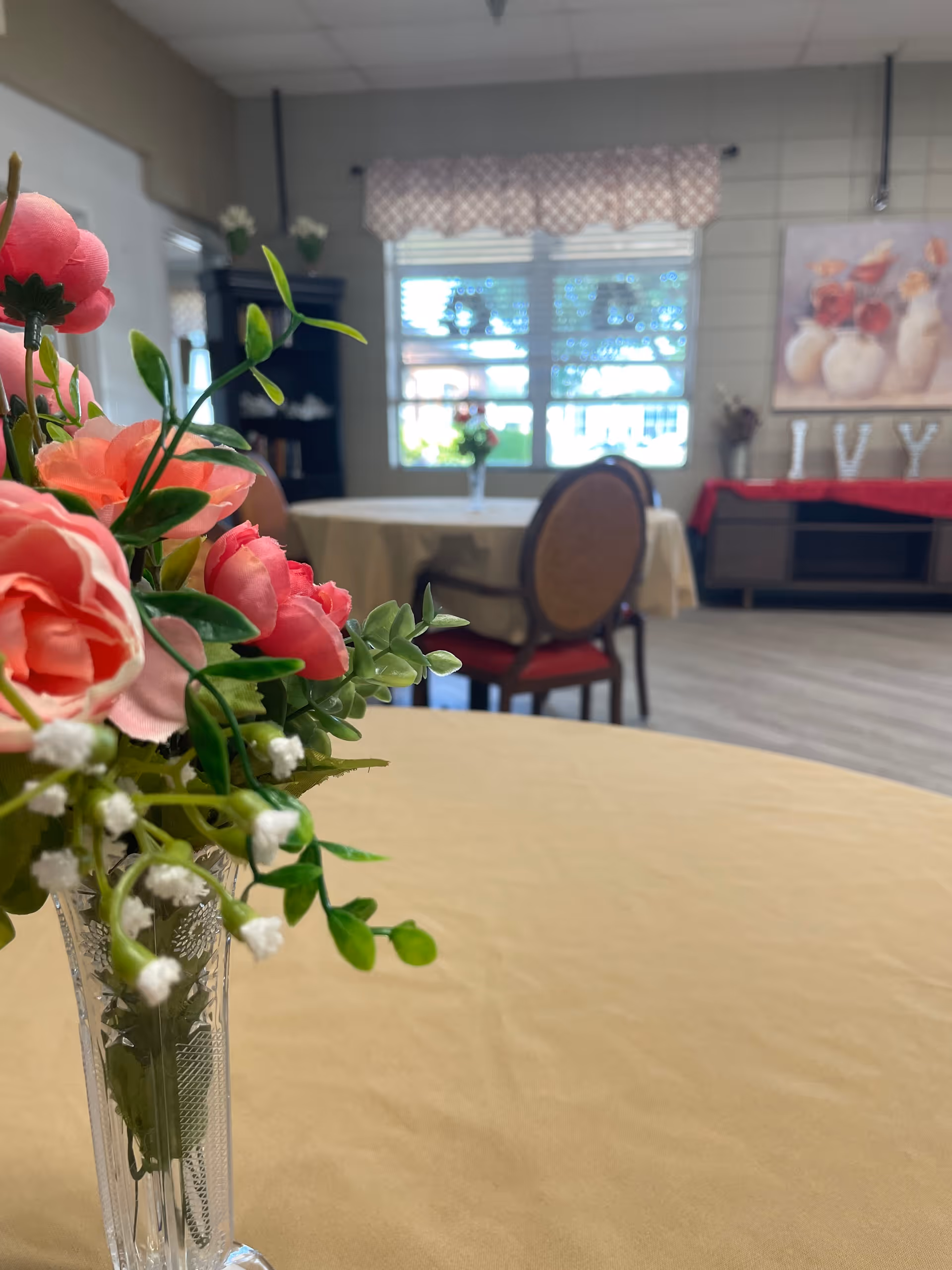 A vase of pink flowers sits on a table in a communal dining/activity room with additional tables, chairs, and a window in the background.