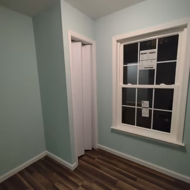 A small empty room with light blue walls, a window with white trim, and a partially open white folding door. The floor is covered with dark wood laminate.