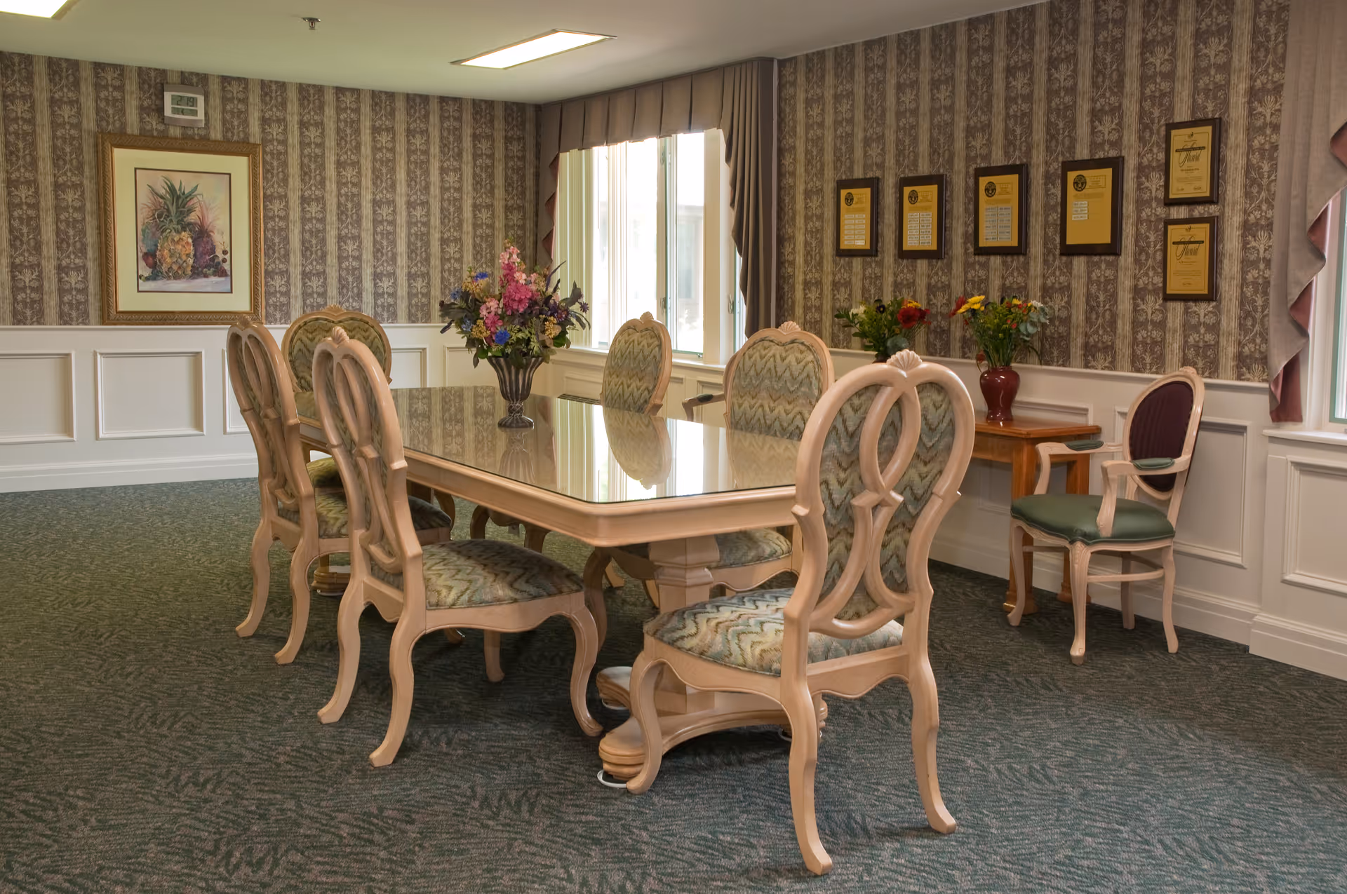A formal dining room with a large rectangular wooden table surrounded by eight upholstered chairs. The room has patterned wallpaper, a carpeted floor, and framed artwork and plaques on the walls. There are two vases with colorful flowers on the table and a small side table.