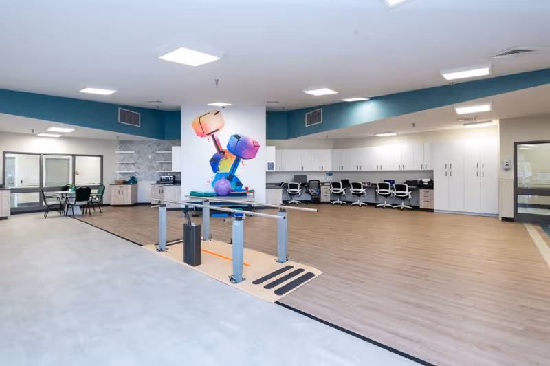 A spacious rehabilitation room with light wood flooring and white walls. The room features a set of parallel bars for physical therapy in the center, a colorful mural of dumbbells on a white pillar, several office chairs lined up against a wall with white cabinets, and a small seating area with tables and chairs near large windows.