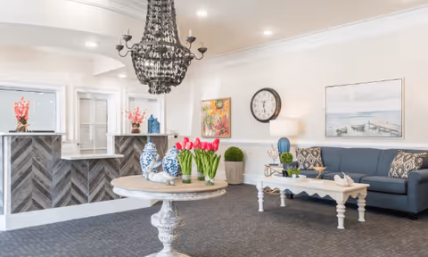 A bright and elegant living room area with a dark blue sofa adorned with patterned cushions, a white coffee table with decorative items, a round table with a vase of pink tulips, and a chandelier hanging from the ceiling. The room features light-colored walls with artwork and a clock, and a reception desk with decorative vases in the background.