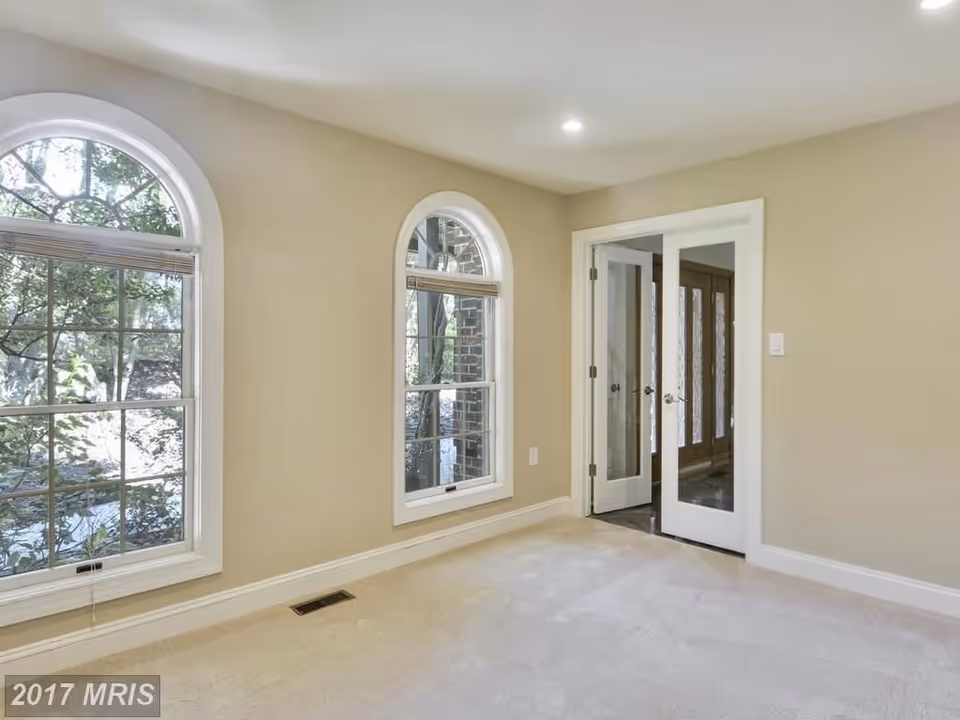 Empty carpeted room with beige walls, two arched windows and glass-paneled double doors.