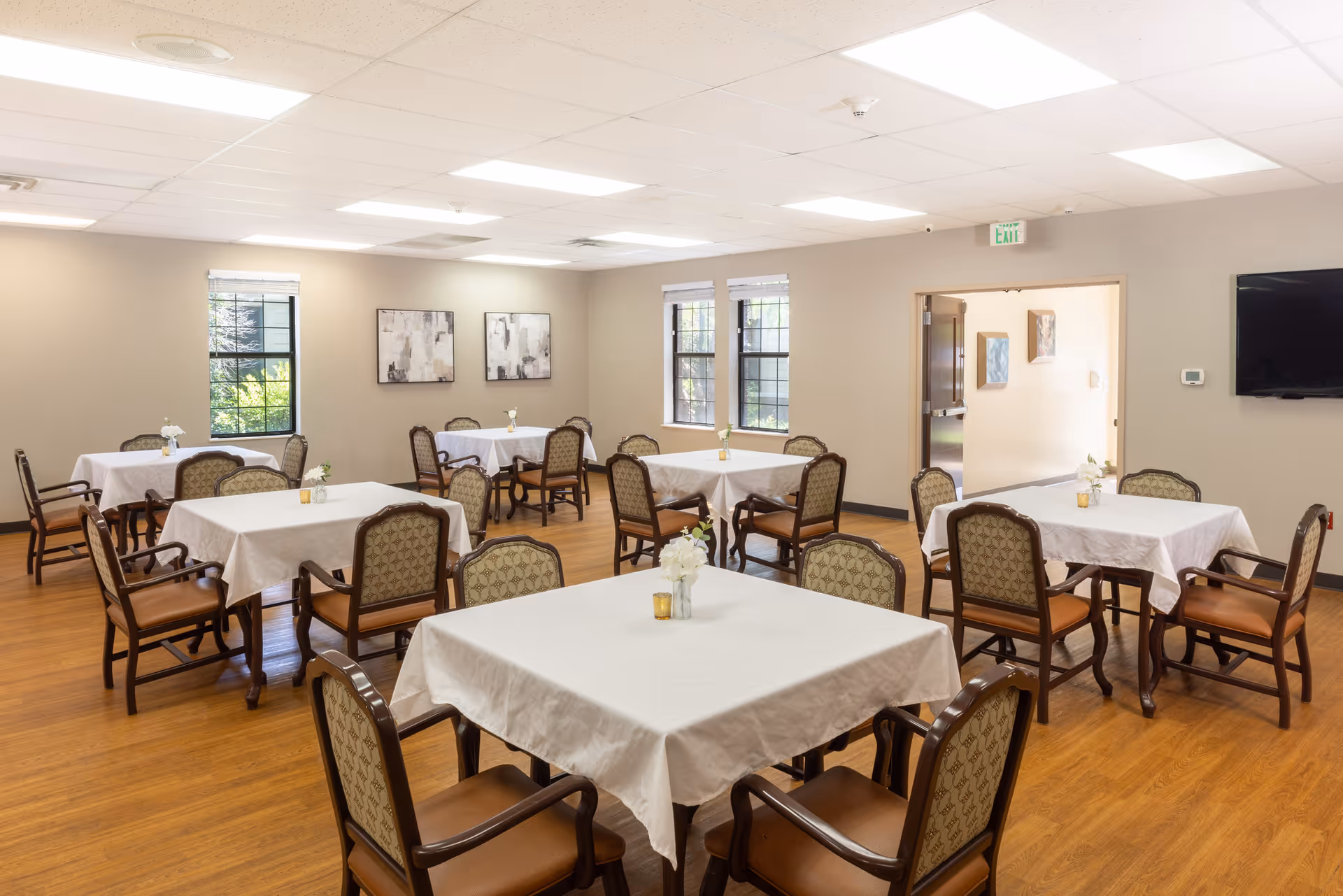 A bright dining room with multiple square tables covered in white tablecloths, each surrounded by four cushioned chairs with wooden frames. Small floral centerpieces and candles are placed on each table. The room has light-colored walls, wooden flooring, several windows letting in natural light, and a wall-mounted television.