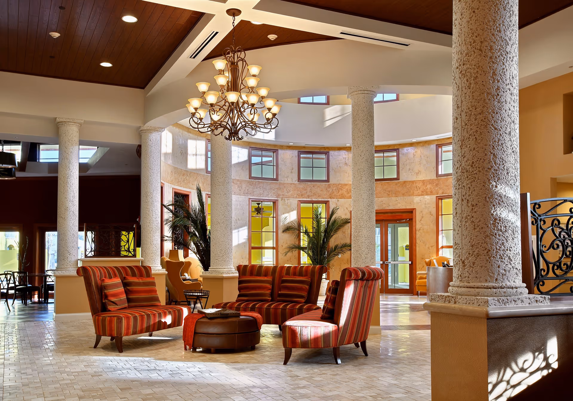 A spacious and elegant living area with high ceilings, large textured columns, and a decorative chandelier. The room features striped upholstered chairs and sofas arranged around a round ottoman. Large windows and glass doors allow natural light to fill the space, and there are potted plants adding greenery to the area.