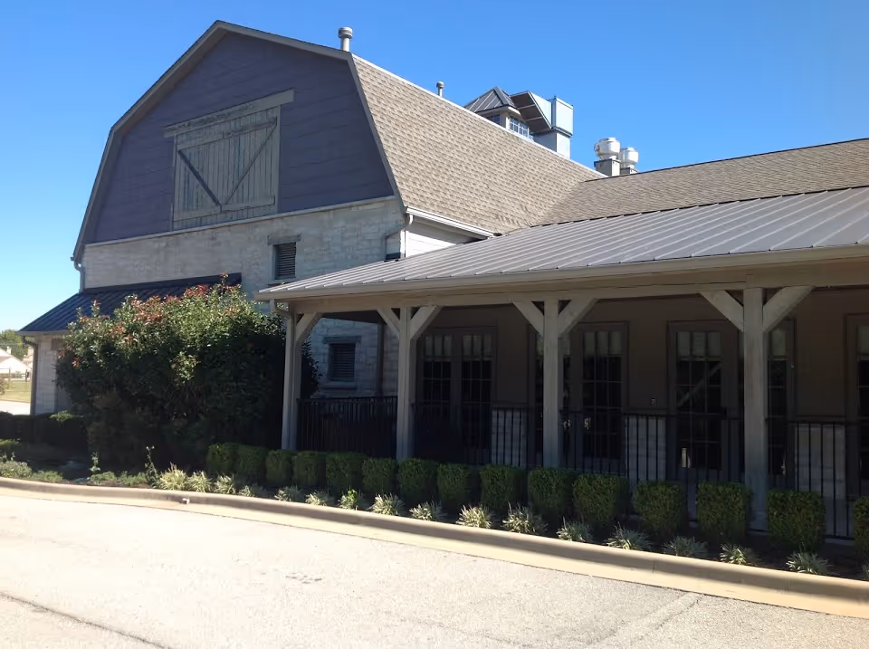 Exterior view of a senior living facility building with a barn-style upper section, stone and wood siding, a covered porch with multiple windows and doors, neatly trimmed bushes, and a clear blue sky.