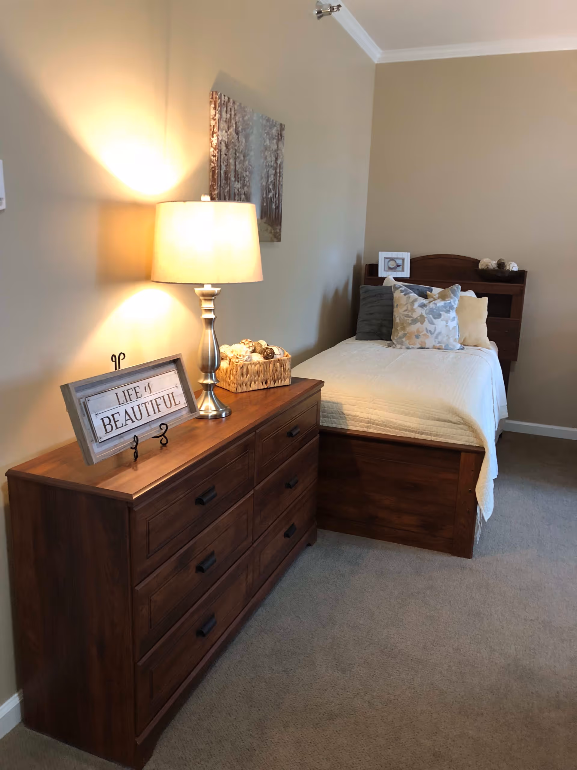 Neat furnished bedroom with a single wooden bed, matching dresser topped with a lamp and a decorative sign that reads 'Life is Beautiful'.