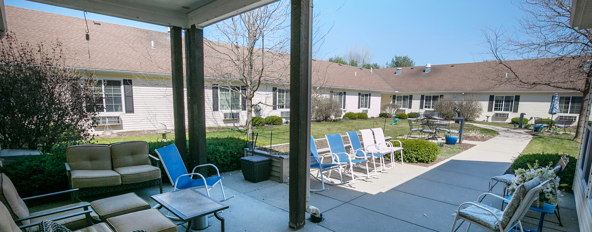 Shaded courtyard with patio seating, lounge chairs, and tables facing a single-story building around a landscaped lawn.