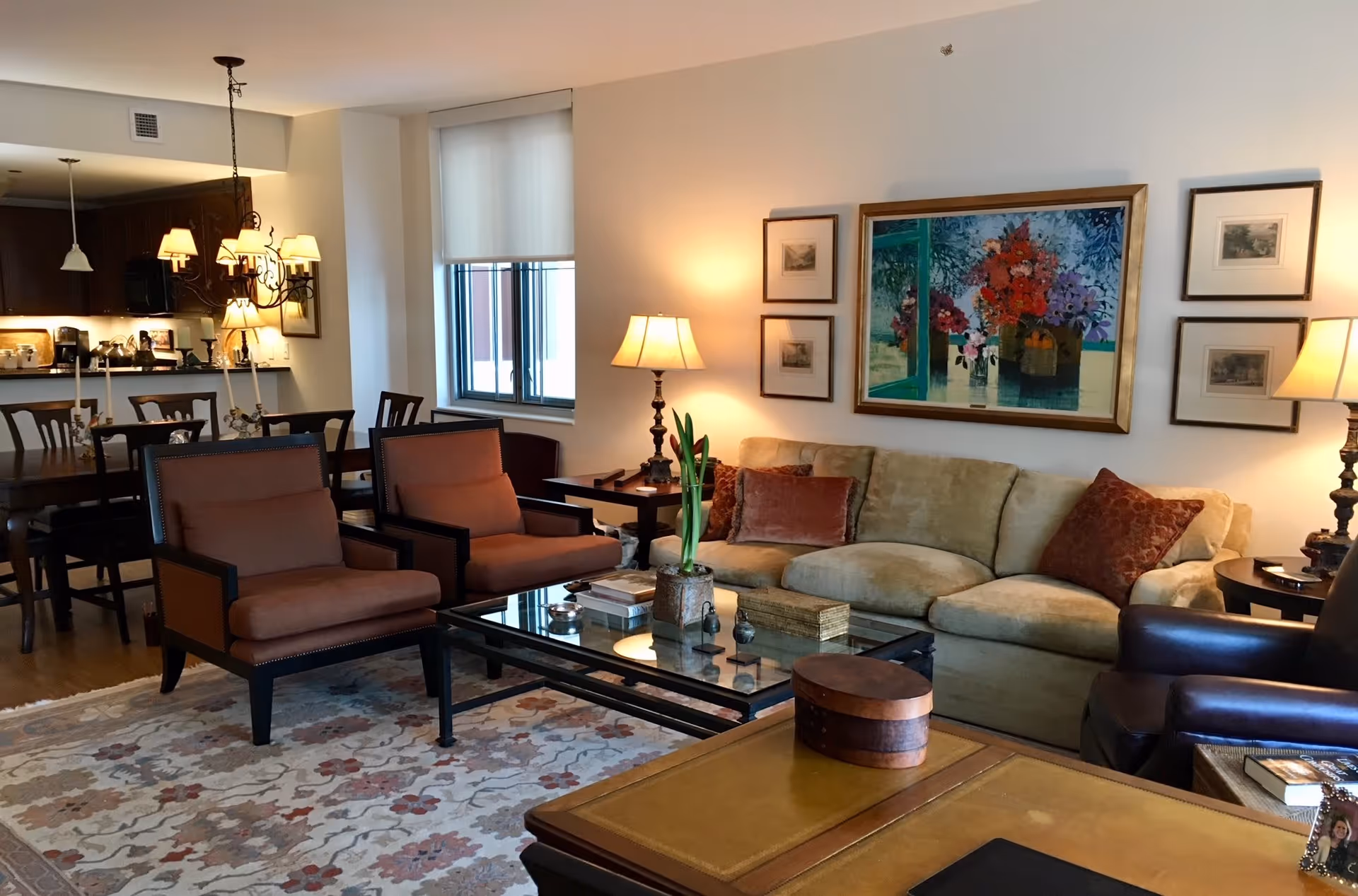 A cozy living room area with a beige sofa adorned with decorative pillows, two brown armchairs, a glass coffee table with books and decorative items, and a patterned rug. The room is warmly lit by table lamps and a chandelier over the adjacent dining table. The walls feature framed artwork, including a large colorful floral painting above the sofa. A window with a white shade lets in natural light, and the background shows a kitchen area with dark cabinetry.
