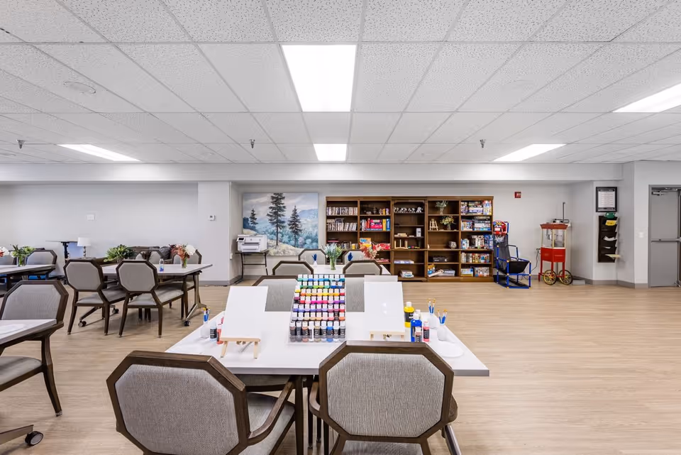 A spacious activity room with tables and chairs arranged for painting and crafts. The tables have easels, paint bottles, and brushes set up. In the background, there is a large bookshelf filled with board games, books, and puzzles. A popcorn machine and a blue chair are visible near the right wall. The room has a light wood floor and a drop ceiling with fluorescent lighting.