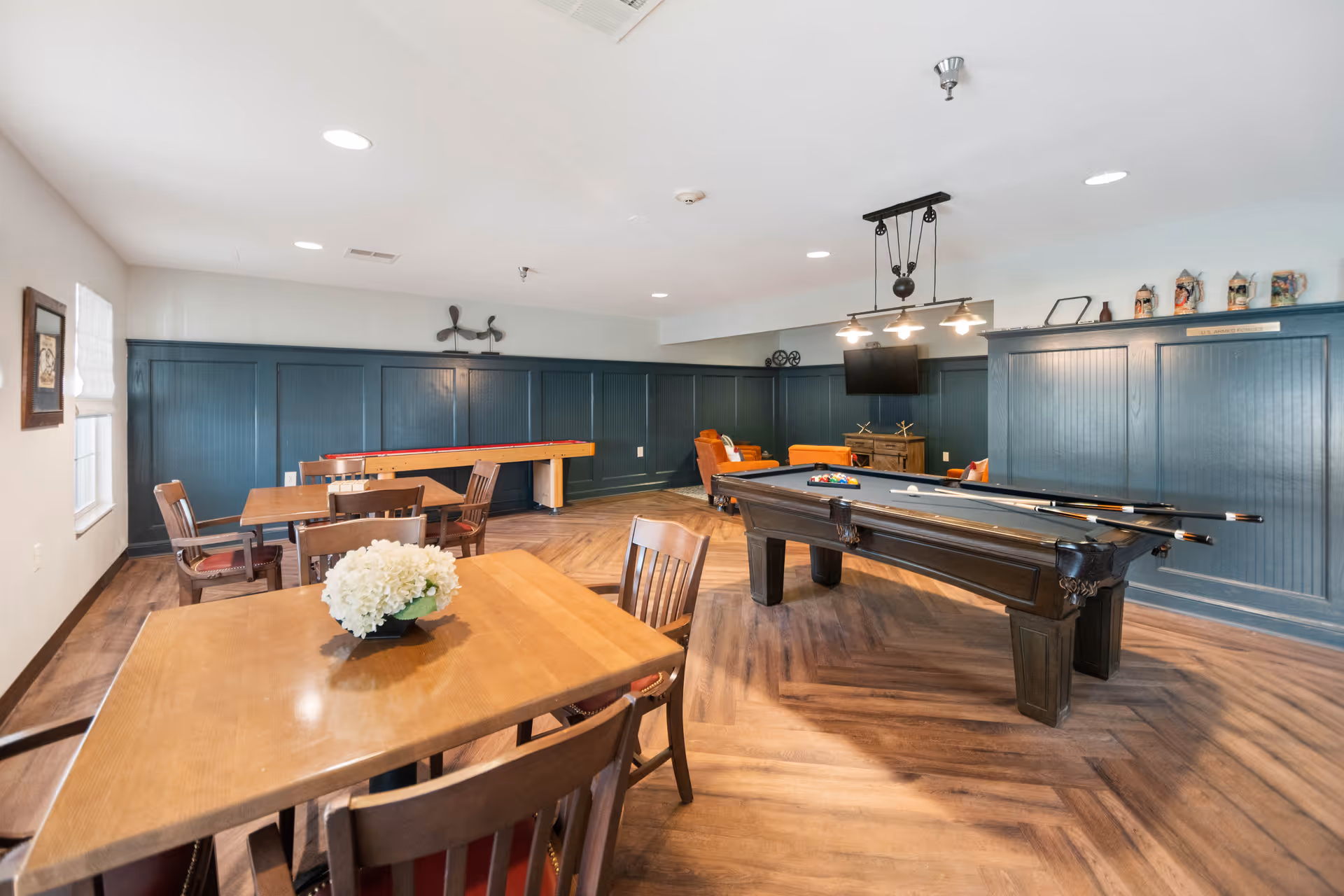 A recreational room with a pool table, shuffleboard table, and several wooden tables and chairs. The room has wood flooring, dark blue wainscoting on the walls, and a mounted TV. There are decorative items on a shelf and a flower arrangement on one of the tables.
