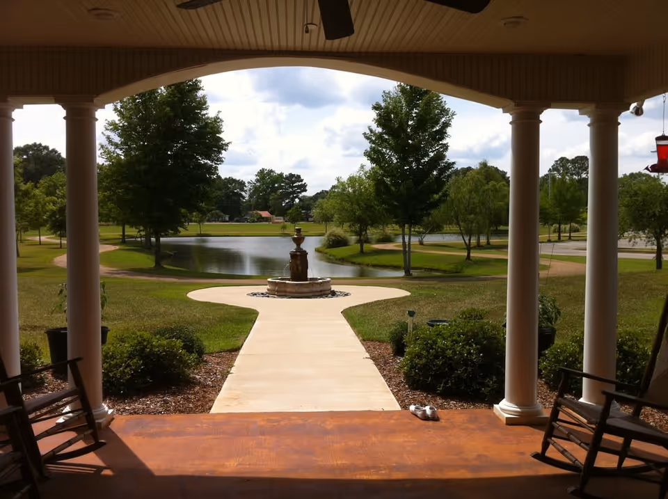 View from a covered porch with white columns and rocking chairs looking out onto a paved walkway leading to a water fountain. Beyond the fountain is a pond surrounded by green grass, trees, and a partly cloudy sky.