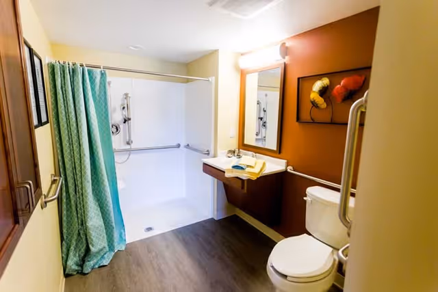 A clean and accessible bathroom with a walk-in shower featuring grab bars and a green shower curtain. There is a wall-mounted sink with a mirror above it, a toilet with grab bars, and a piece of wall art with abstract shapes on a brown accent wall.