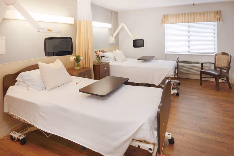 A bright and clean room with two hospital-style beds, each with white linens, pillows, and an adjustable overbed table. There are small bedside tables with flower vases between the beds. Mounted on the wall above each bed is a television screen attached to an adjustable arm. A window with blinds and a valance lets in natural light. A wooden chair is placed near the window on a wood floor.