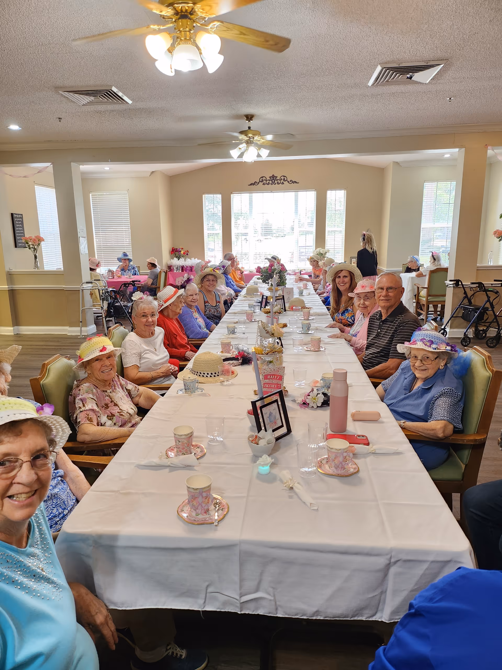 A group of elderly people sitting around a long dining table in a well-lit room, wearing colorful hats and smiling. The table is set with teacups, saucers, and small decorations. The room has large windows letting in natural light, ceiling fans, and a warm, inviting atmosphere.