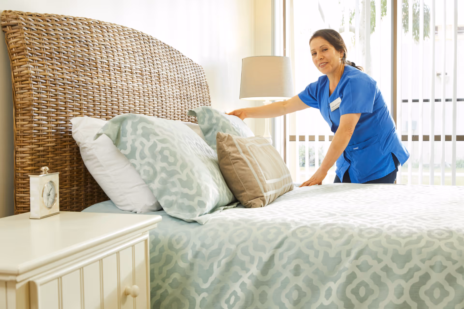 A woman in blue scrubs is making a bed with a wicker headboard in a bright room with a window and a lamp on a nightstand.