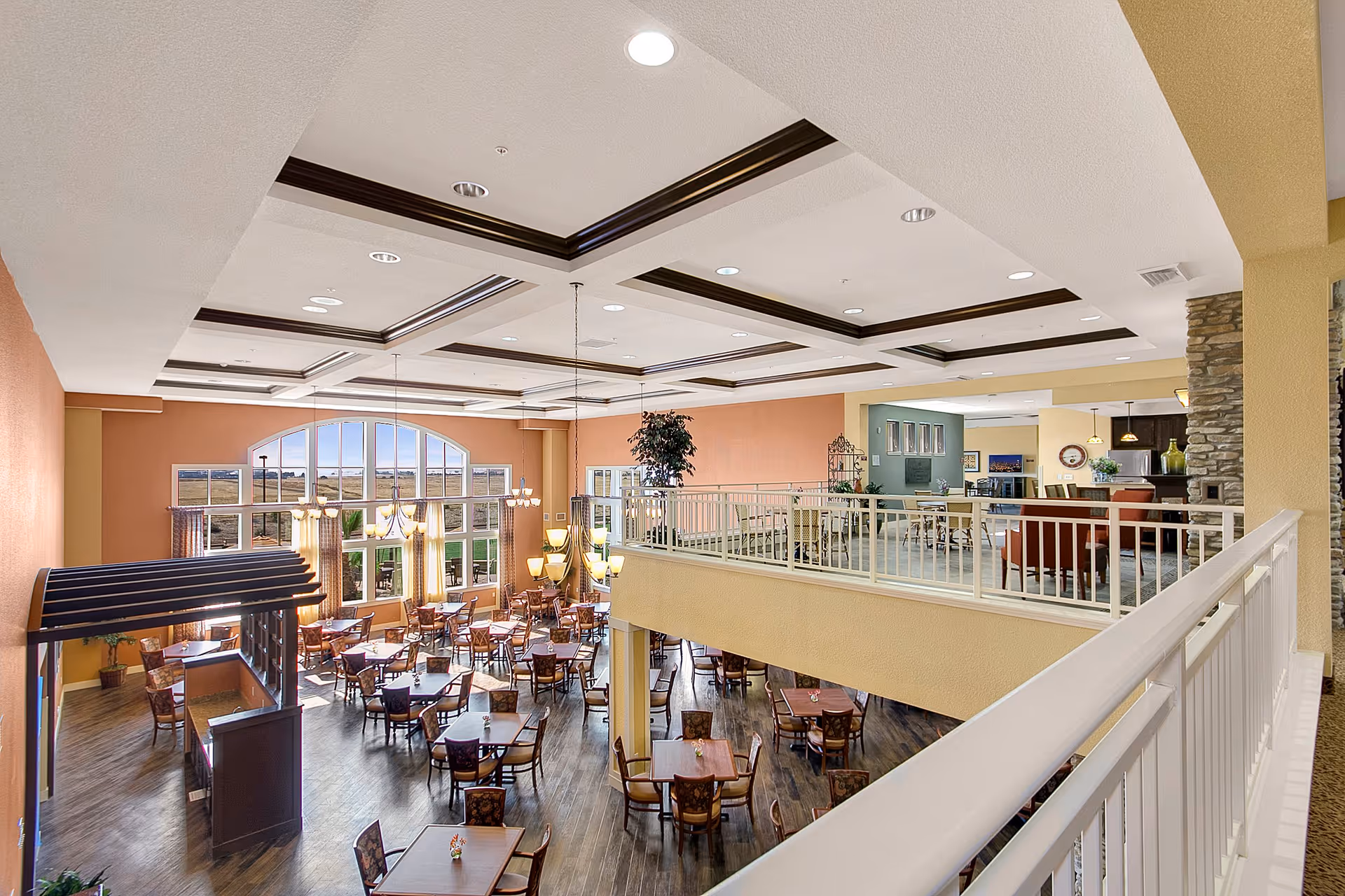 Spacious dining area in a senior living facility with multiple wooden tables and chairs arranged neatly on a dark wood floor. Large arched windows allow natural light to fill the room. The ceiling features recessed lighting and decorative beams. An upper level with additional seating and a stone fireplace is visible, separated by a white railing.