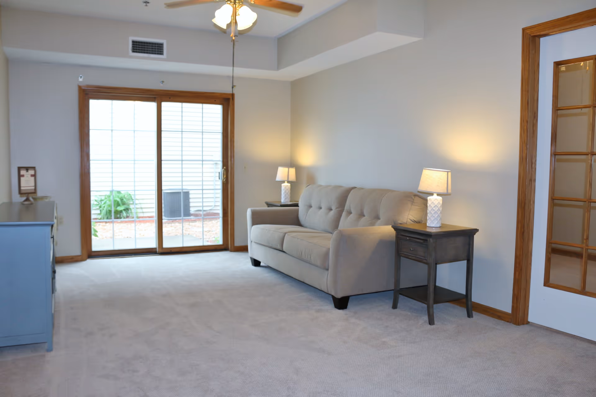 A living room with a beige sofa flanked by two dark wooden side tables, each with a white lamp. There is a ceiling fan with lights above, a blue dresser on the left, and a sliding glass door with wooden trim leading outside. The room has light-colored carpet and neutral walls.