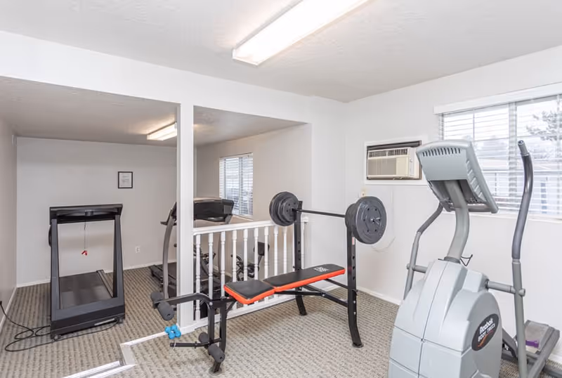 Carpeted fitness room with a treadmill, weight bench and elliptical trainer near windows and a wall air conditioner.