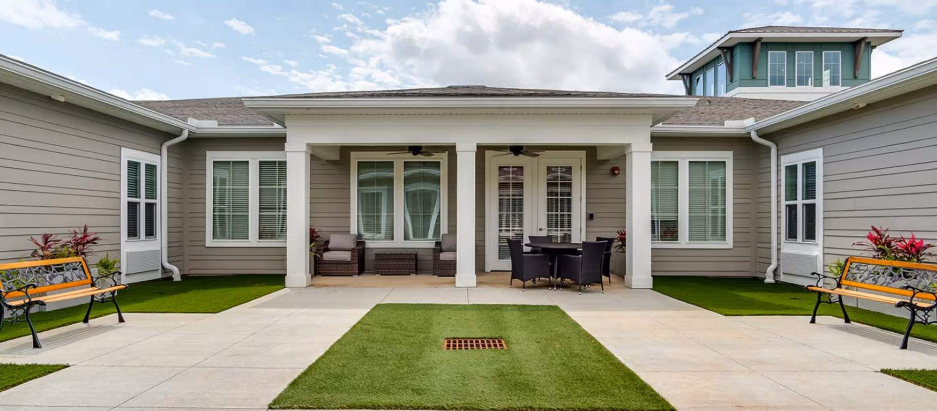 Outdoor courtyard area of The Canopy at Duval Station featuring two wooden benches with black metal frames on either side, a central concrete walkway with a rectangular patch of green grass, and a covered patio with wicker chairs and a round table with chairs. The building exterior is light gray with white trim and multiple windows.