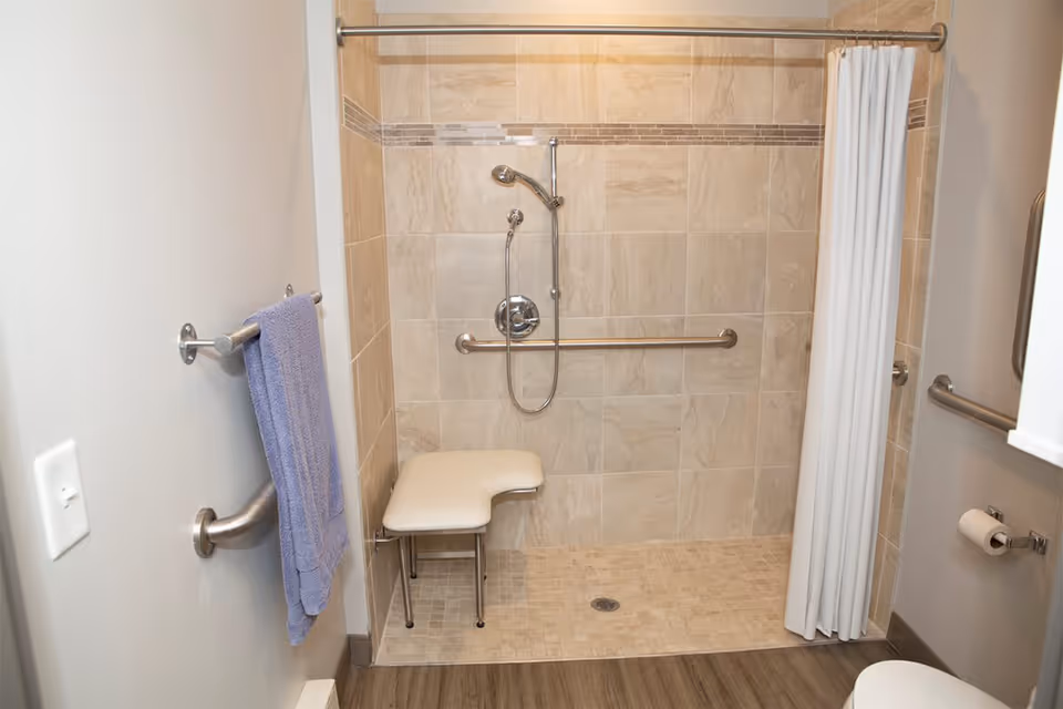Accessible shower area with beige tiled walls and floor, a white shower seat, a handheld showerhead, grab bars on the walls, a white shower curtain, a towel rack with a blue towel, and a toilet paper holder.