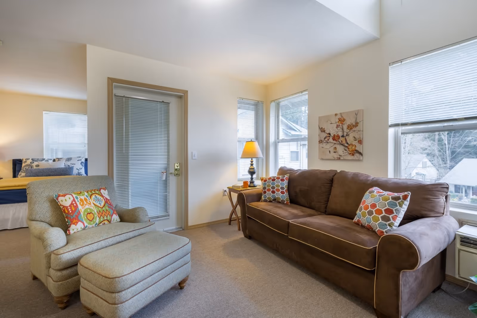 A cozy living room area in a senior living facility featuring a brown sofa with colorful hexagonal patterned pillows, a beige armchair with a matching ottoman and a colorful pillow, a side table with a lamp, and windows with blinds letting in natural light. A bed is partially visible in an adjacent room.