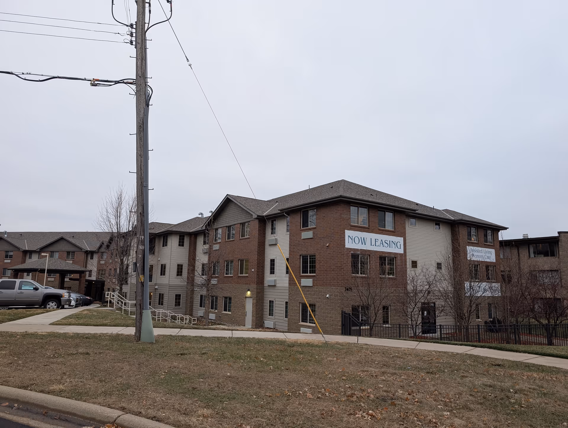 Exterior view of a multi-story senior living facility building with brick and siding exterior. Signs on the building read 'NOW LEASING' and 'Assisted Living Memory Care'. There is a sidewalk, some parked cars, and a utility pole in the foreground under an overcast sky.