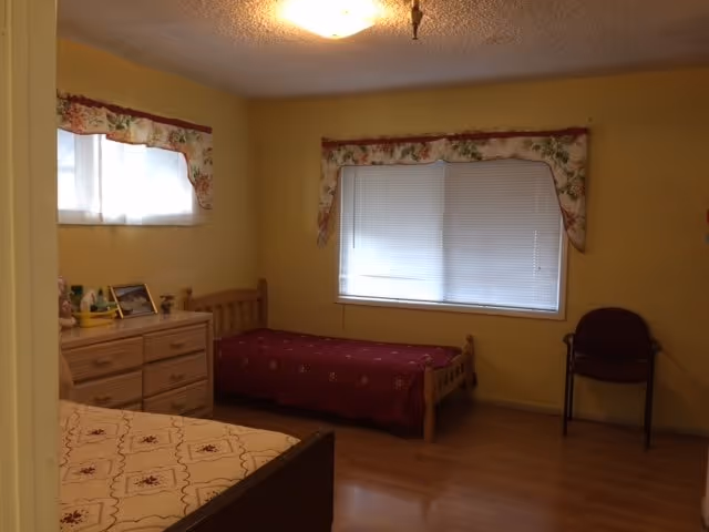A simple bedroom with two single wooden beds, one with a red bedspread and the other with a beige patterned bedspread. There is a wooden dresser with framed photos and small decorative items on top. The room has yellow walls, two windows with floral valances, and a single chair near the window. The floor is wooden.