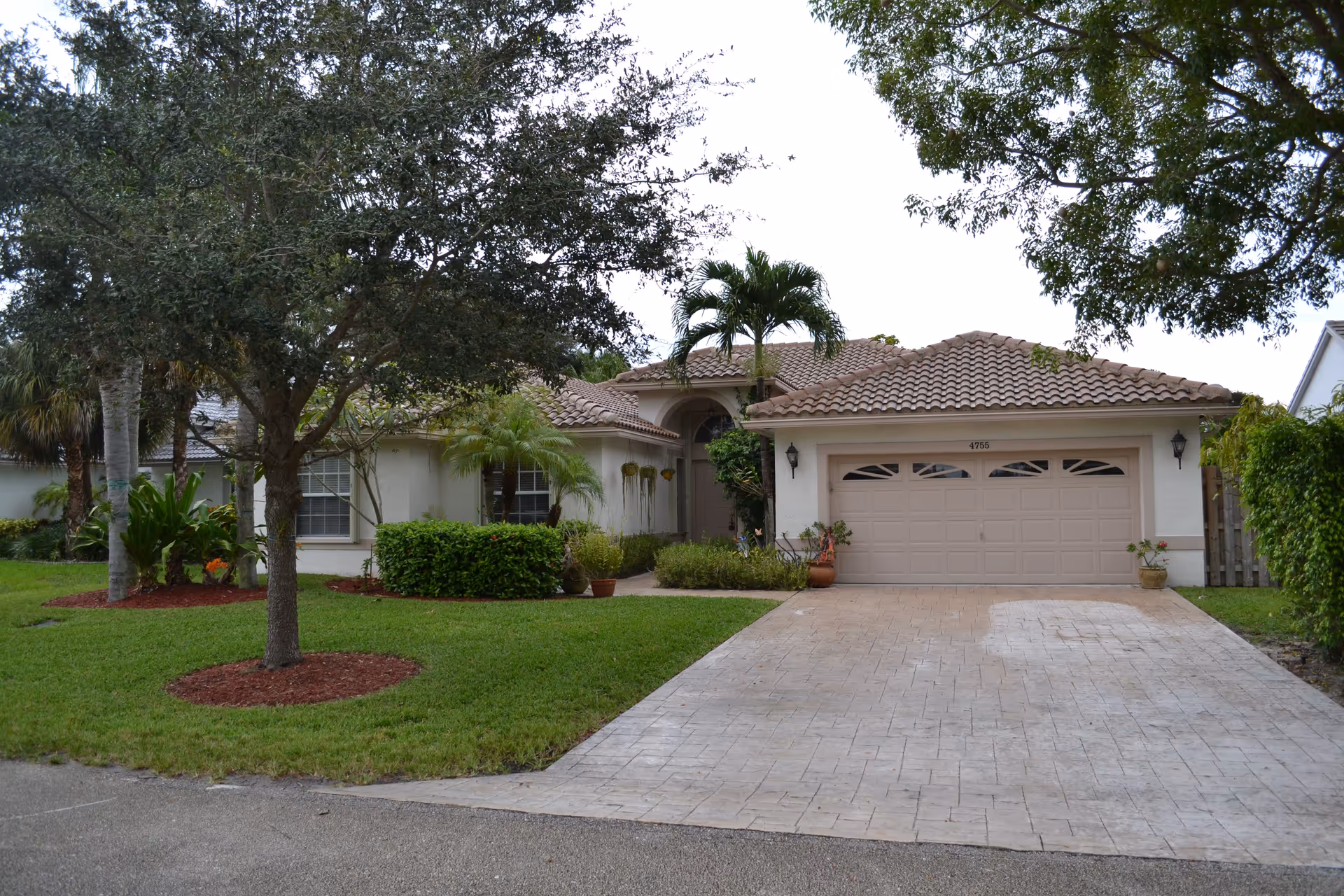 Front exterior view of a single-story house with a tiled roof, a two-car garage, a driveway, and a well-maintained lawn with trees and shrubs.