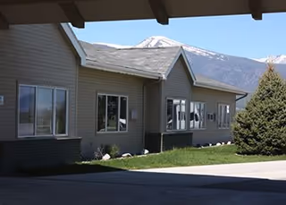 Single-story senior living building exterior with multiple windows, a covered driveway, lawn and a snow-capped mountain in the background.