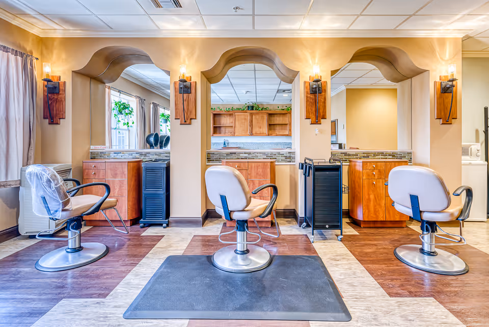 Interior view of a salon area with three salon chairs in front of three large mirrors mounted on a beige wall. Each station has wooden cabinetry and wall-mounted light fixtures. The floor has a patterned design with different shades of brown and beige. There are windows with curtains on the left side and some plants visible through the mirrors.
