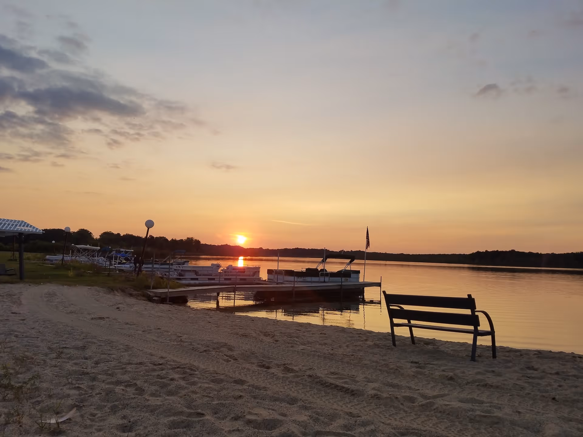 Sunset view over a calm lake with several boats docked at a pier. A sandy beach with a single bench facing the water is in the foreground. The sky is partly cloudy with warm colors from the setting sun.