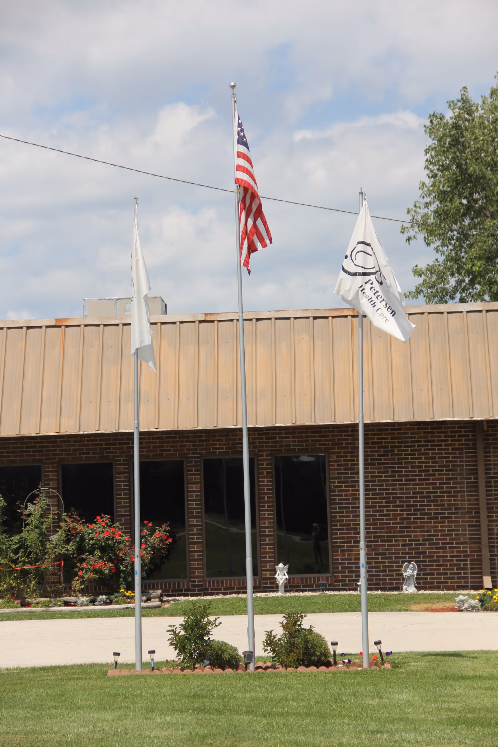 Three flagpoles with flags in front of a brick building. The middle flagpole displays the American flag, while the other two have white flags. There are small bushes and flowers planted around the base of the flagpoles, and the building has large dark windows. The sky is partly cloudy.