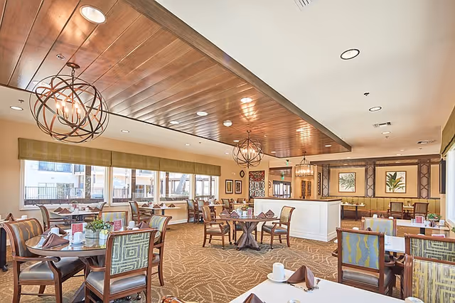 Well-lit dining room with round and rectangular tables, upholstered chairs, decorative chandeliers, and large windows.