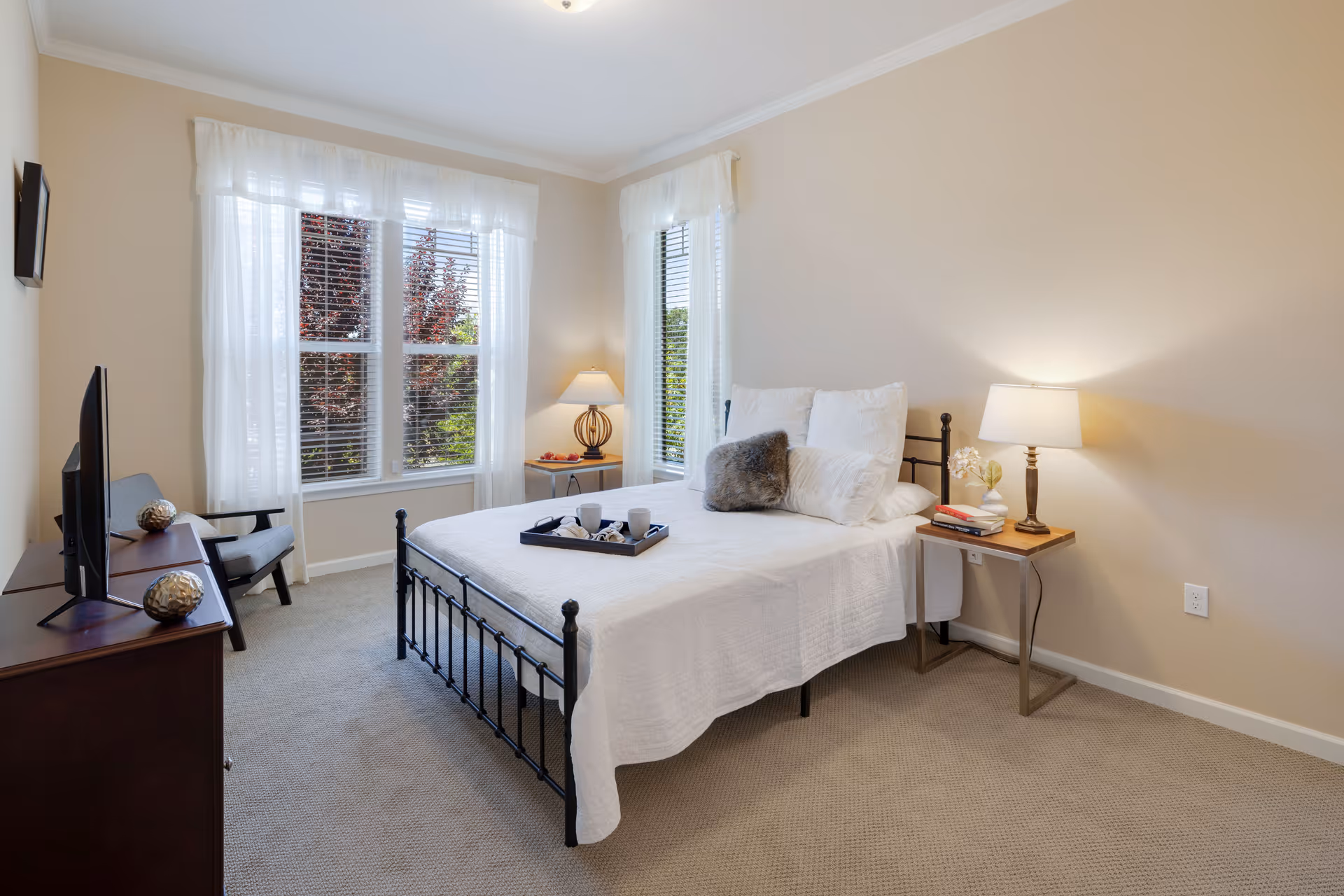 A bright bedroom with beige walls and carpet, featuring a black metal bed frame with white bedding and multiple pillows. There are two bedside tables with lamps, a tray with cups on the bed, a TV on a wooden dresser, and a gray armchair near large windows with white sheer curtains letting in natural light.