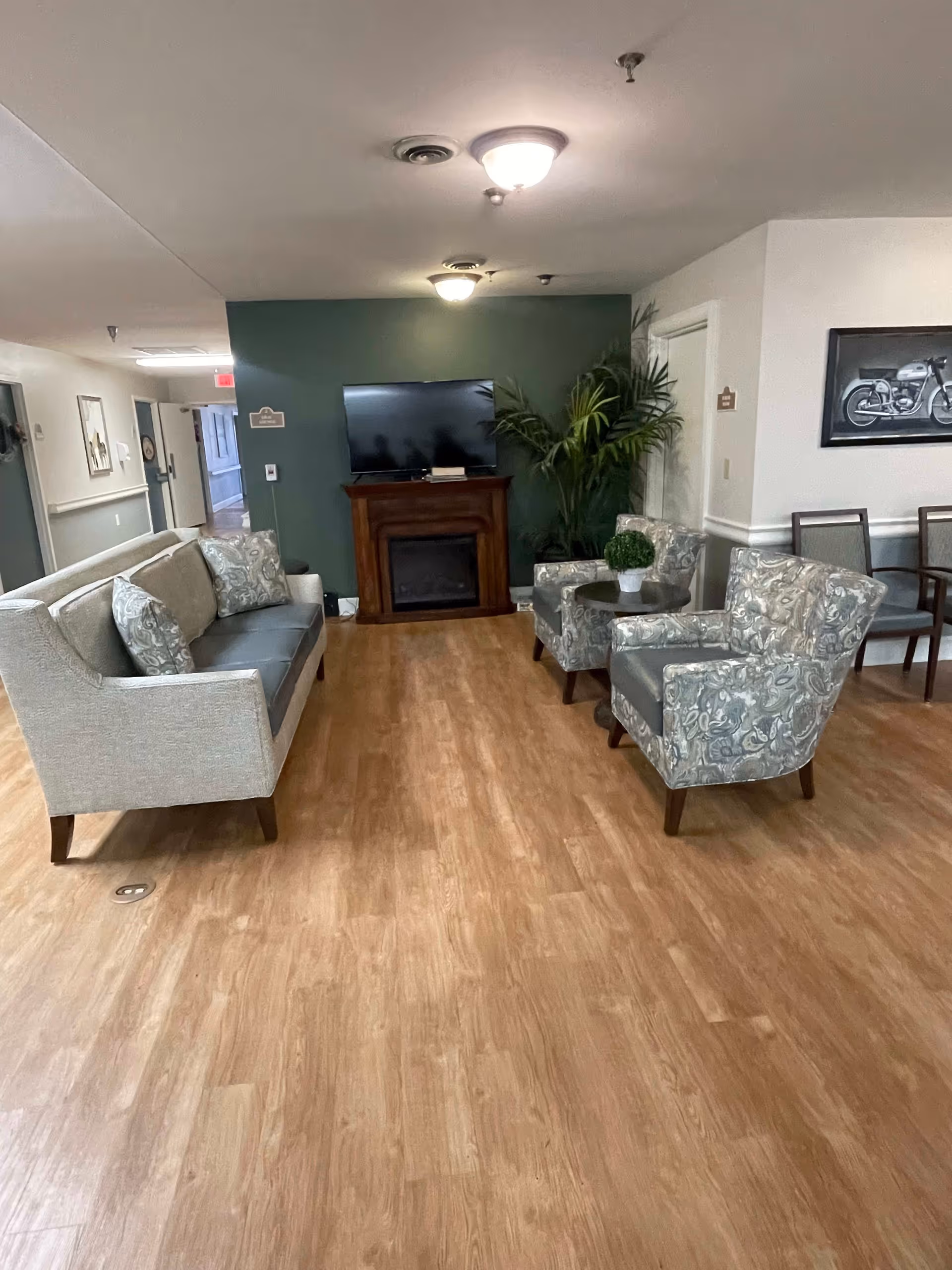 A cozy living room area in a senior living facility with a gray sofa and two patterned armchairs arranged around a small round table with a potted plant. A flat-screen TV is mounted above a wooden fireplace against a green accent wall. There are additional chairs along the right wall and a framed picture of a motorcycle hanging above them. The floor is wood laminate, and the ceiling has two light fixtures.