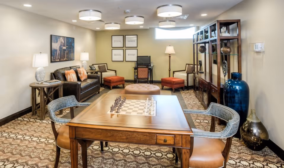 Well-appointed lounge with a wooden chess table and chairs in the foreground, a seating area with sofa, armchairs, ottomans, shelving and decorative vases.