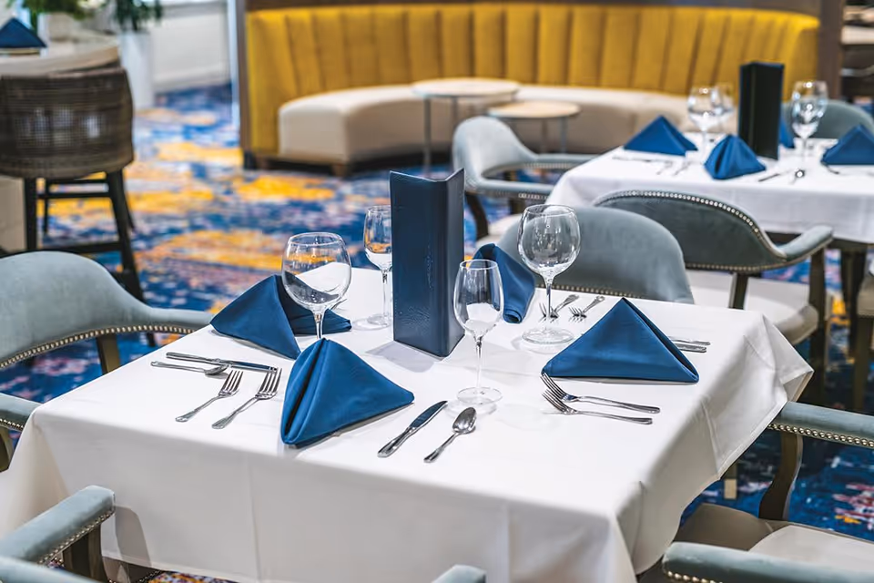 A dining table set with white tablecloth, blue folded napkins, wine glasses, and silverware in a senior living facility dining room with upholstered chairs and a yellow curved booth in the background.