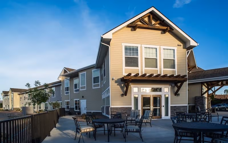 Exterior of a two-story senior living building with a paved patio area furnished with tables and chairs under a clear blue sky.