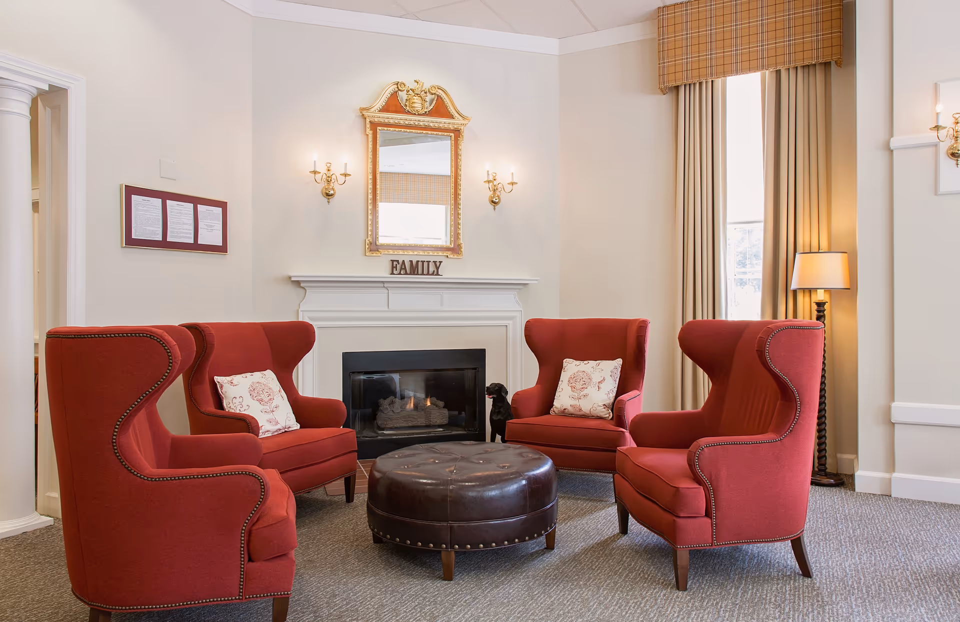 A cozy sitting area with four red wingback chairs arranged around a round brown leather ottoman. Behind the chairs is a white fireplace with a gold-framed mirror above it and the word FAMILY displayed on the mantel. The room has beige walls, a window with beige curtains and a plaid valance, and a floor lamp in the corner.