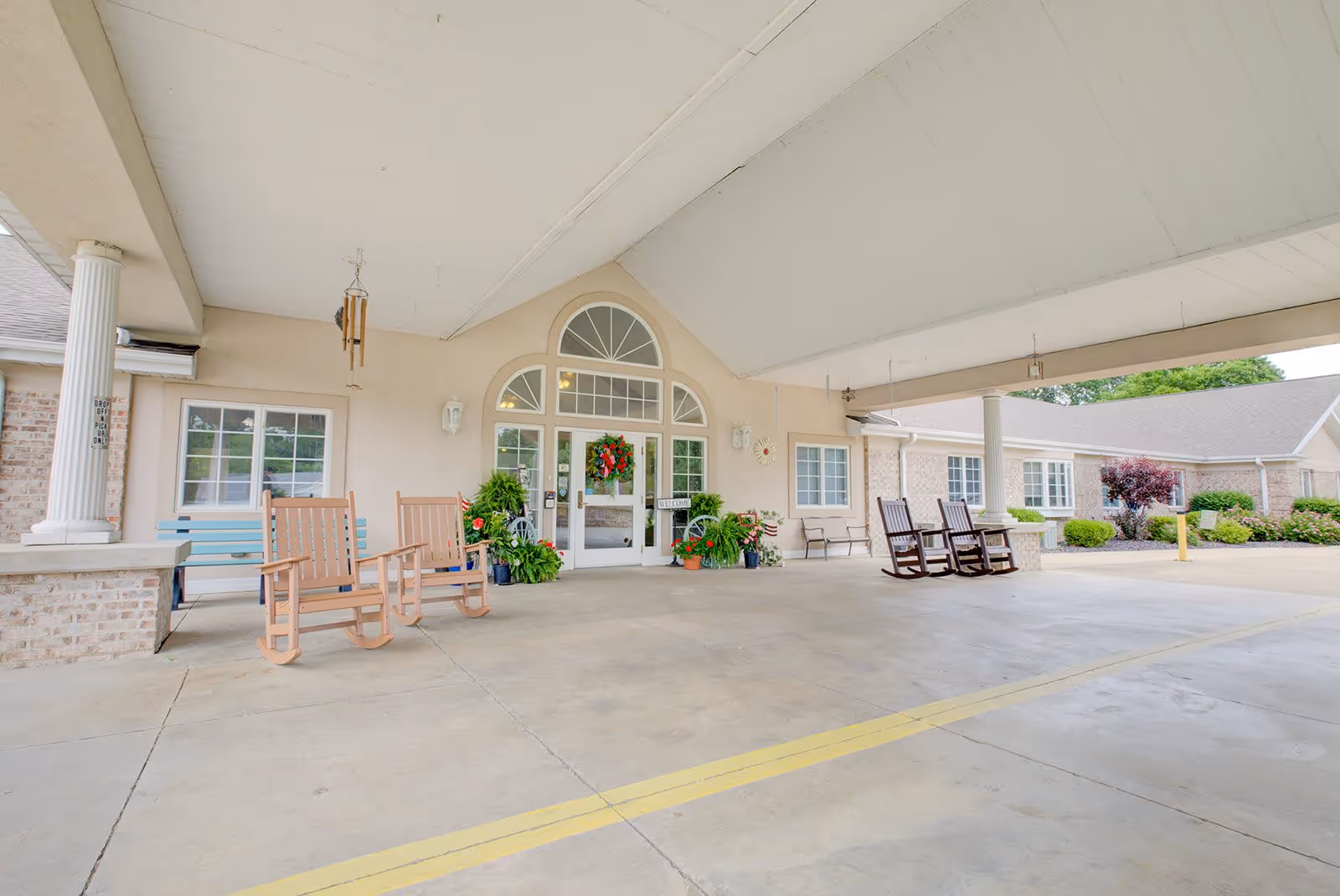 Covered entrance area of Brookstone Estates of Vandalia featuring a wide driveway with yellow lines, several rocking chairs, potted plants, and a glass door entrance decorated with a wreath. The building exterior is light-colored with brick accents and white columns supporting the roof.