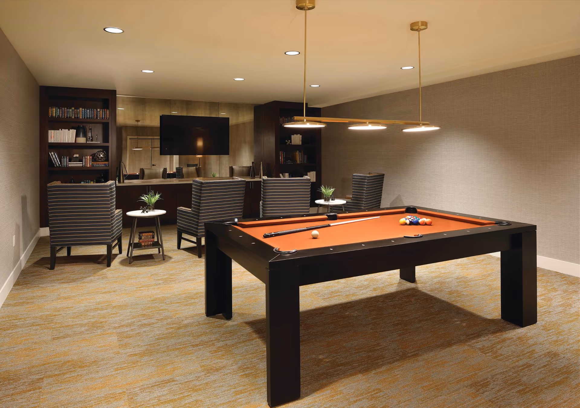 A well-lit recreational room featuring a pool table with an orange felt surface and pool balls arranged on it. Behind the pool table are four striped armchairs arranged around two small round tables with potted plants. There are built-in dark wood bookshelves filled with books and decorative items, and a large flat-screen TV mounted on the wall above a long dark wood cabinet. The room has beige walls and carpeted flooring with a subtle pattern.