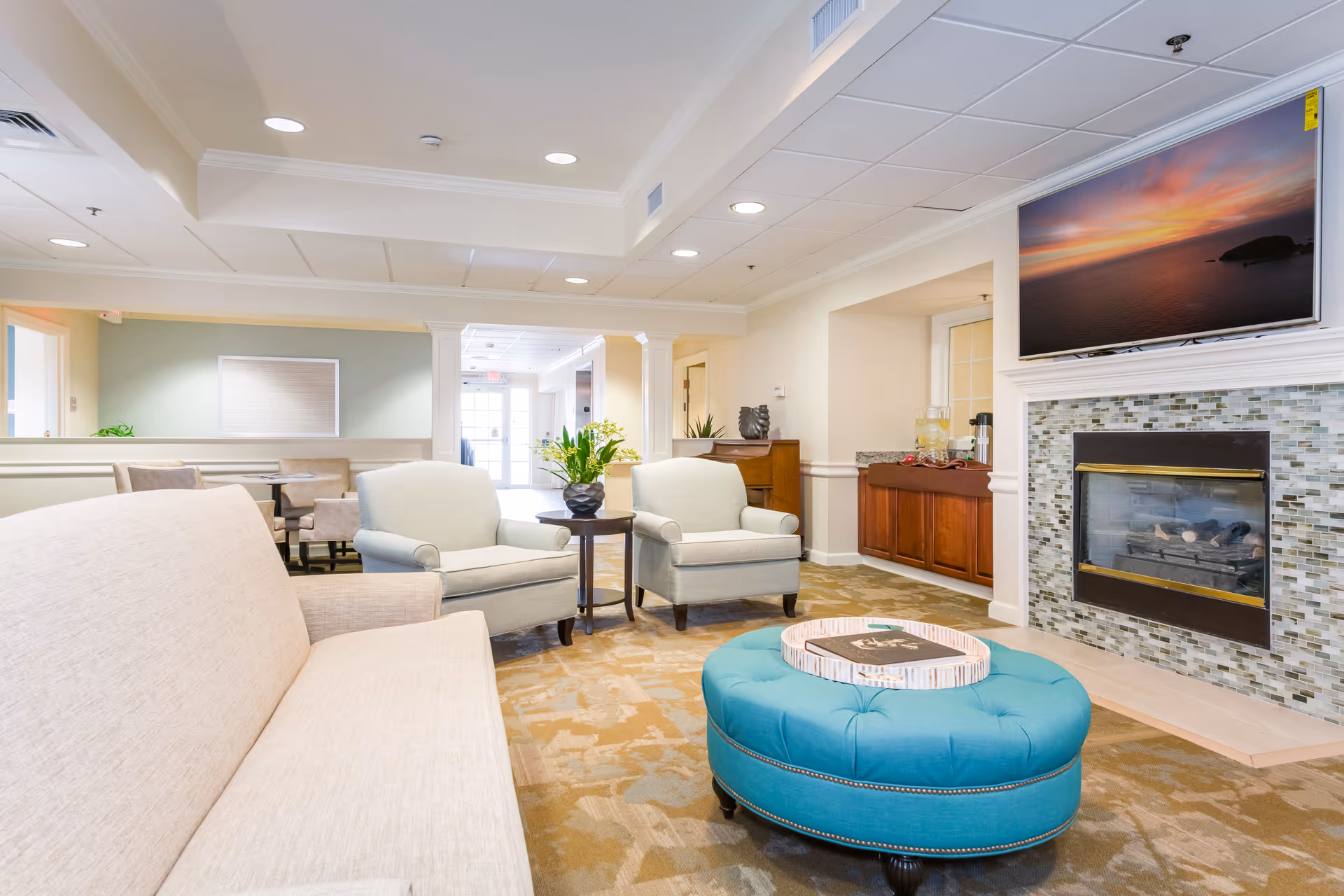 A bright and inviting senior living facility lounge area with beige and light blue armchairs, a beige sofa, a round turquoise ottoman with a tray on top, a fireplace with a mosaic tile surround, and a large flat-screen TV mounted above the fireplace displaying a sunset over the ocean. There is a small table with a plant between the armchairs and a wooden cabinet with refreshments in the background.