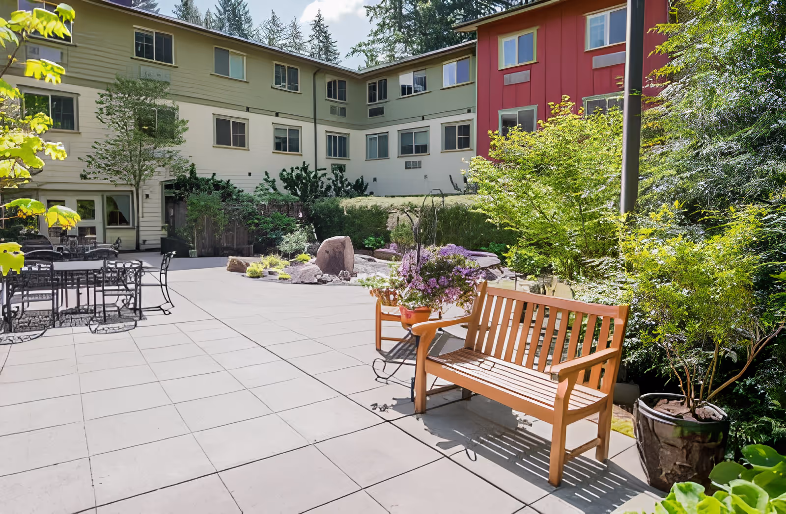 Outdoor patio area at Village Concepts of Issaquah-Spiritwood at Pine Lake featuring a wooden bench, potted plants, metal tables and chairs, surrounded by greenery and a multi-story building in the background.