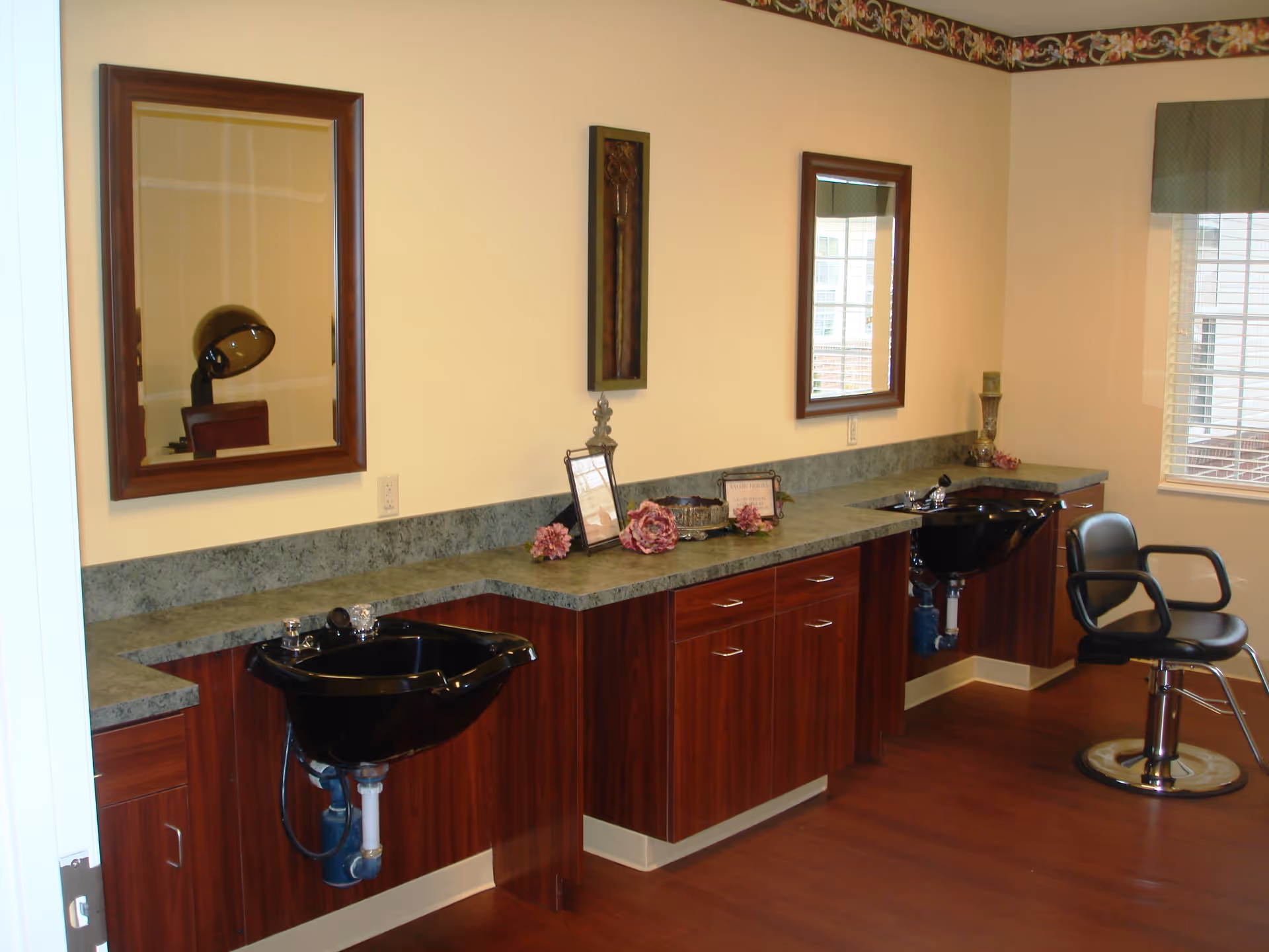Interior view of a hair salon area in a senior living facility with two black hair washing sinks, wooden cabinets with green countertops, two mirrors mounted on the wall, a salon chair, and decorative items on the countertop. A window with blinds and a valance is visible on the right side.