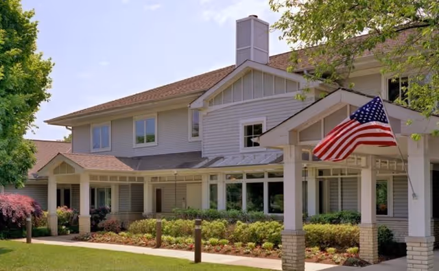 Exterior view of a two-story senior living facility building with light gray siding, a brown shingled roof, and a covered entrance displaying an American flag. The surrounding area includes well-maintained landscaping with bushes, flowers, and trees under a clear blue sky.
