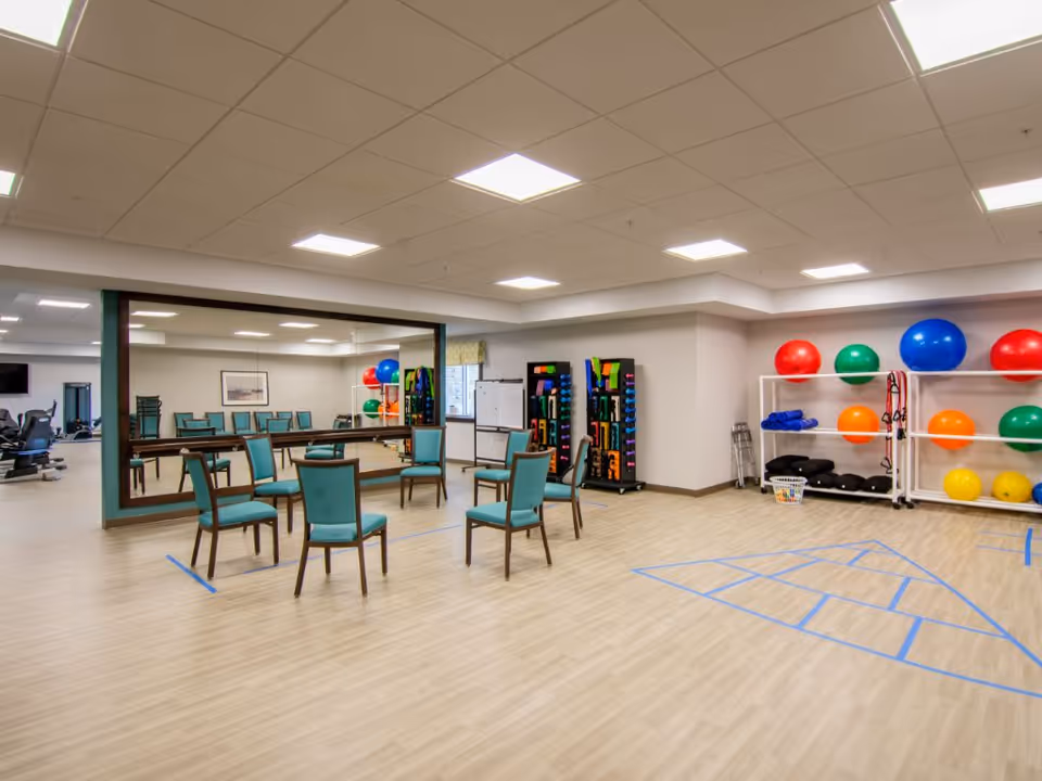 A spacious exercise room with wooden flooring and a large mirror on one wall. The room contains several teal chairs arranged in a circle, exercise equipment including colorful exercise balls on white racks, resistance bands, and weights. There is a whiteboard and a window with a patterned valance. The ceiling has recessed lighting.
