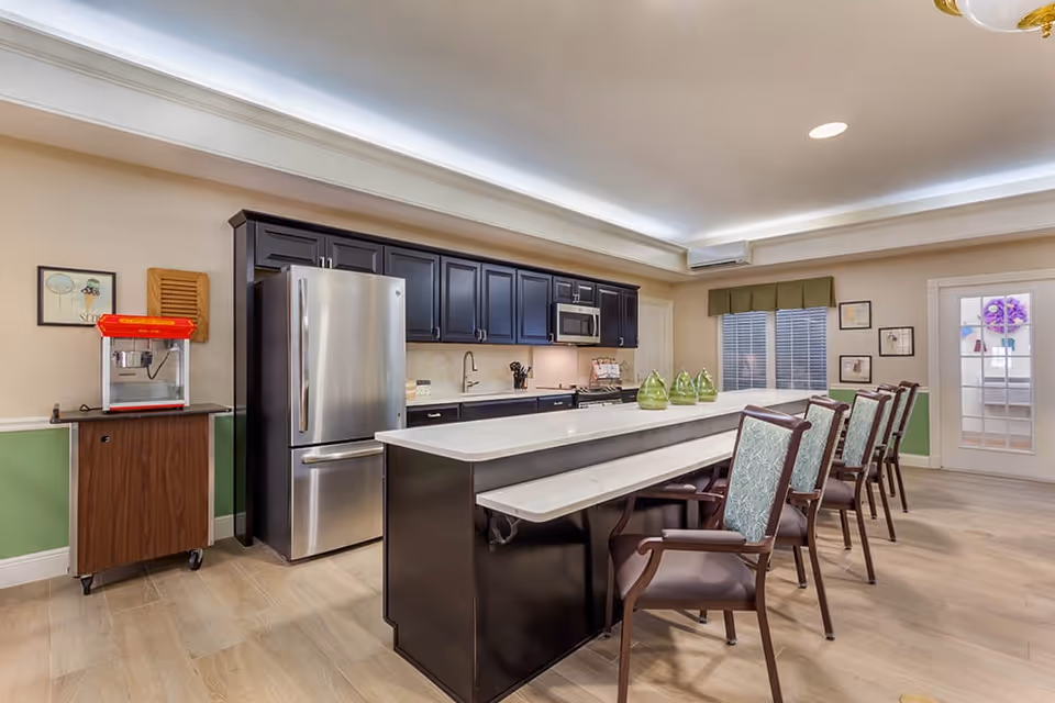A modern kitchen area with dark cabinetry, a stainless steel refrigerator, microwave, and a long white countertop island with several chairs lined up along one side. There is a popcorn machine on a small wooden cart to the left, light wood flooring, and a window with a green valance. The room has soft beige walls with a green lower half and framed pictures on the walls.