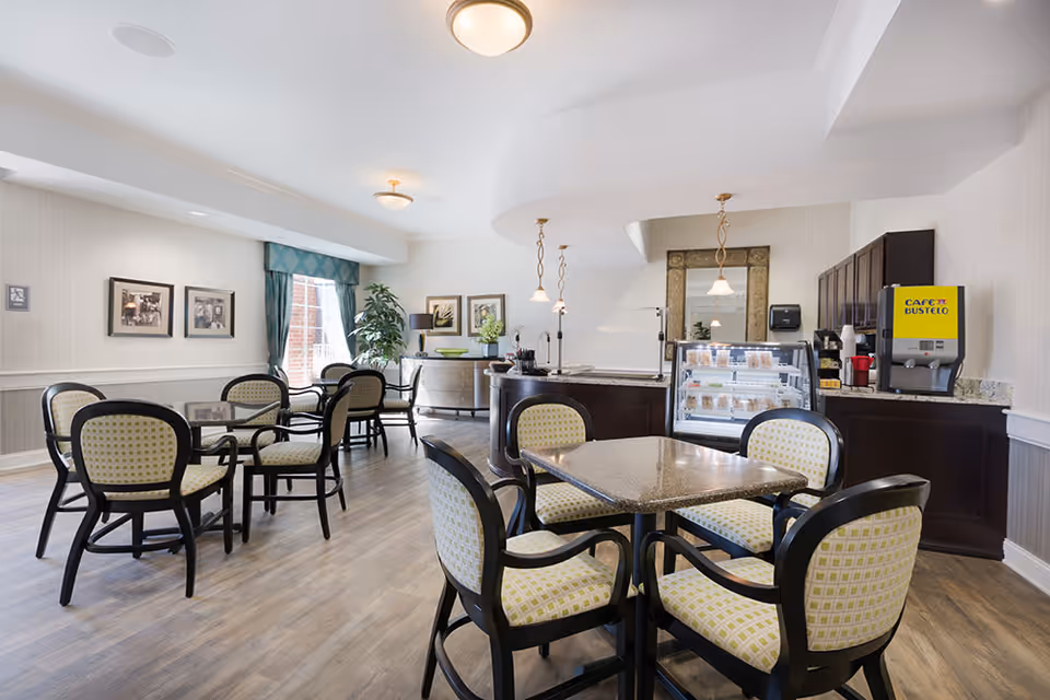 A bright and clean dining area with several tables and chairs featuring patterned upholstery. The room has light-colored walls, wood flooring, framed pictures on the wall, and a large window with green curtains letting in natural light. In the background, there is a counter with a glass display case containing food items and a Café Bustelo coffee machine.