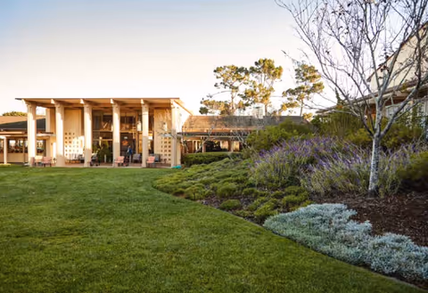 Outdoor view of Canterbury Woods facility showing a well-maintained lawn, landscaped garden with various shrubs and flowers, and a building with large windows and a covered patio area under a clear sky.