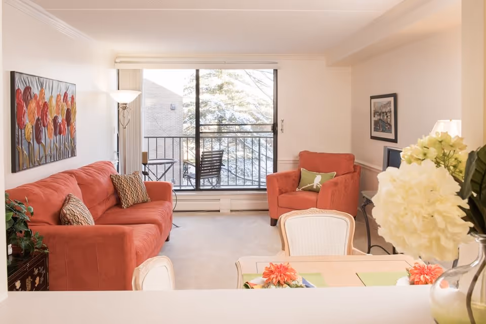 A bright living room with a large window and sliding glass door leading to a balcony. The room features a red sofa with patterned pillows, a matching red armchair with a green pillow, a floor lamp, a painting of colorful flowers on the wall, and a small table with a lamp and framed picture. In the foreground, there is a dining table with chairs and a vase of white flowers.